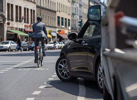Radfahrer in dunkler Kleidung mit Helm fährt auf einer Stadtstraße, im Vordergrund steht ein schwarzes Auto am Straßenrand, weitere Fahrzeuge und Gebäude im Hintergrund.