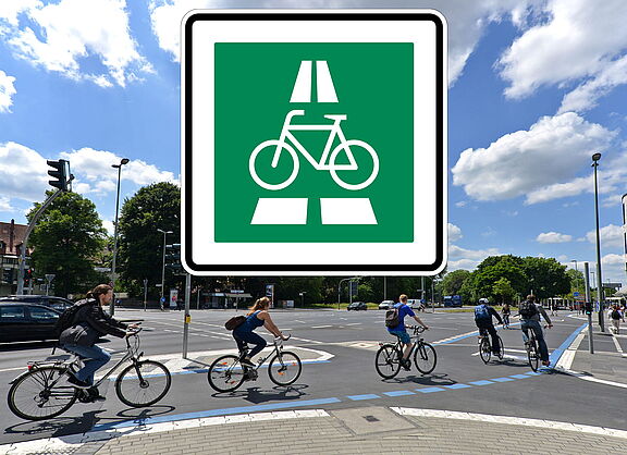 Quadratisches, grünes Verkehrsschild mit weißem Rand. In der Mitte ist ein weißes Fahrrad-Symbol abgebildet. Darüber und darunter deuten weiße Balken eine Fahrbahn an. Im Hintergrund fahren mehrere Personen auf Fahrrädern über eine Kreuzung.