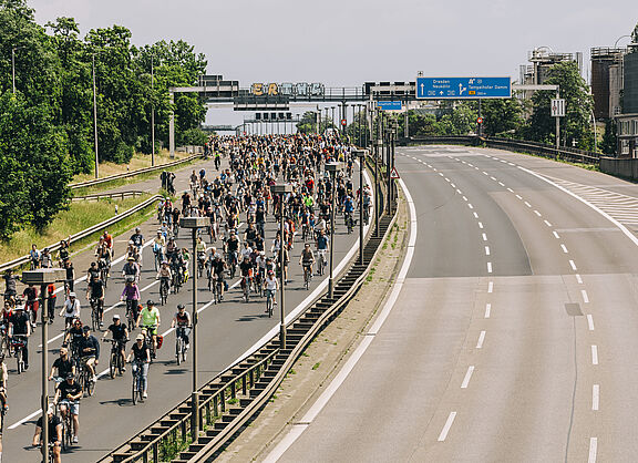 Hunderte Radfahrende bei der Berliner Sternfahrt auf einer gesperrten Autobahn. Die Teilnehmenden fahren gemeinsam auf der linken Fahrbahn, während die rechte Seite leer bleibt. Im Hintergrund sind Autobahnbrücken und Verkehrsschilder zu sehen.